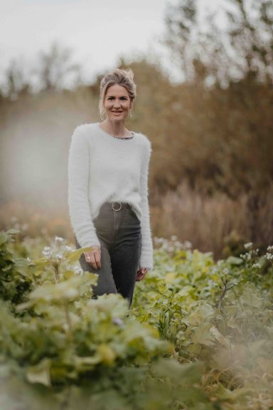 Junge Frau in einem Garten steht zwischen Pflanzen und lächelt in die Kamera.