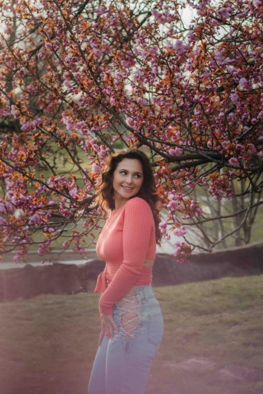 Eine lächelnde Frau in einem rosa Pullover steht vor blühenden Kirschbäumen.