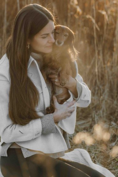 Eine Frau mit langen Haaren hält einen kleinen Hund in einem Feld bei Sonnenuntergang.