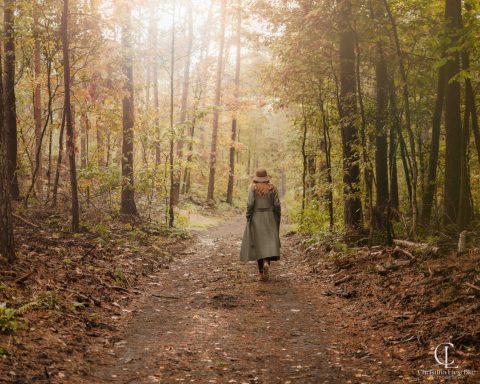 Frau in langen Kleidern geht einen Waldweg entlang, umgeben von herbstlichen Bäumen.