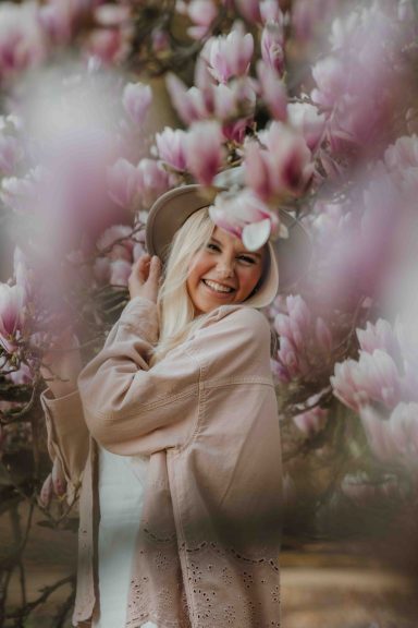 Eine lachende Frau mit blonden Haaren steht unter Magnolienblüten.