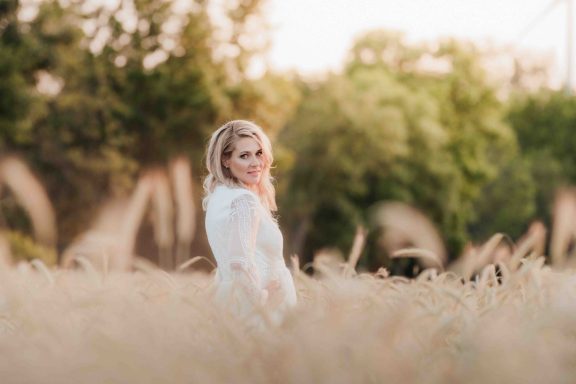 Junge Frau in einem weißen Kleid steht lächelnd in einem goldenen Feld.
