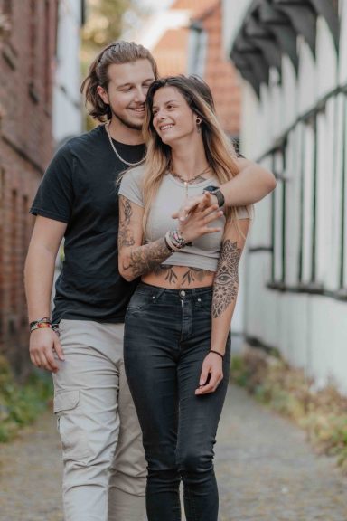 Ein Paar mit Tattoos, das entspannt auf einem Weg neben einer Mauer steht und lächelt.