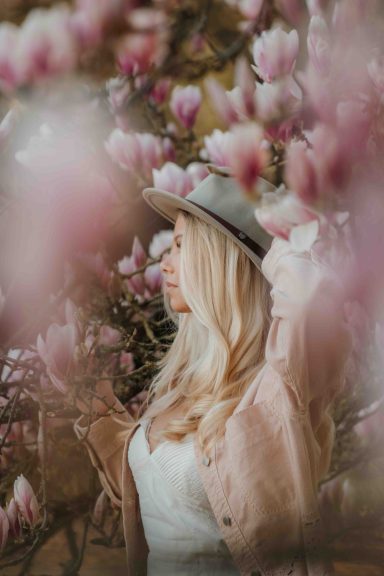 Frau mit langen blonden Haaren und Hut vor blühenden Magnolien.