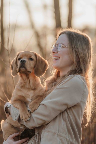 Frau mit Brille hält einen Hund im Freien, umgeben von sonnigem, natürlichem Licht.
