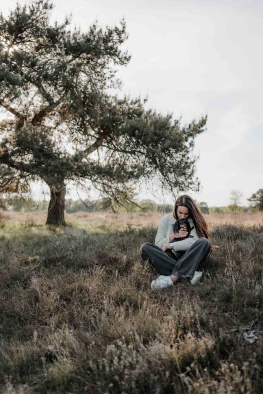 Junge Frau sitzt im Freien auf dem Boden und liest ein Buch neben einem Baum.