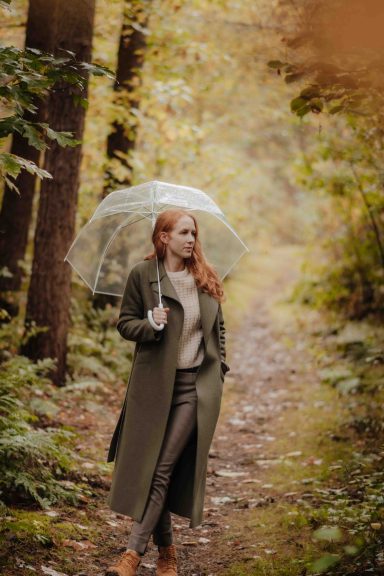 Frau mit rotem Haar trägt einen Mantel und hält einen Regenschirm in einem Wald.