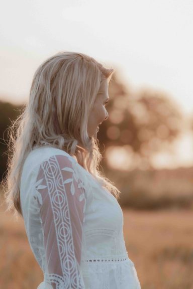 Frau in weißem Kleid, steht in einem Feld und blickt nachdenklich zur Seite.