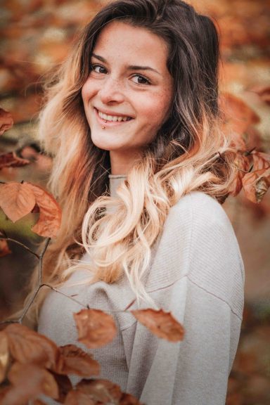 Junge Frau mit langen, gewellten Haaren, umgeben von herbstlichem Laub.