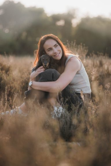 Frau umarmt einen Hund in einem Feld mit sanftem Licht im Hintergrund.