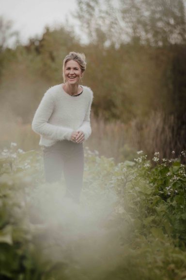 Eine lächelnde Frau steht im Grünen, umgeben von Blumen und sanfter Nebelatmosphäre.