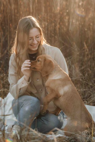 Eine lächelnde Frau sitzt auf einer Decke und interagiert mit einem Hund im Freien.