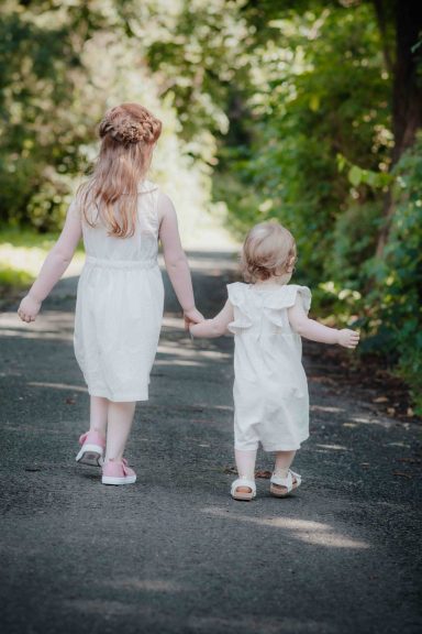 Zwei Mädchen in weißen Kleidern, Hand in Hand, gehen einen Weg entlang.