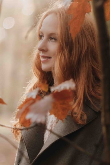 Eine lächelnde Frau mit langen roten Haaren, umgeben von herbstlichen Blättern.