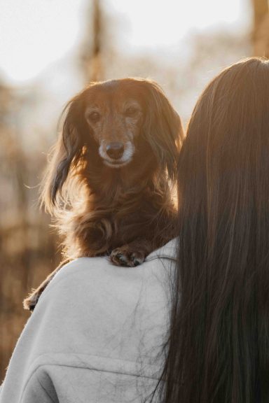 Frau mit langen Haaren trägt einen kleinen Hund auf der Schulter.